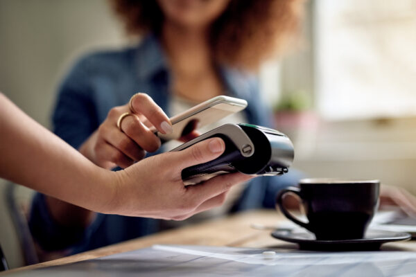 A person holds a smartphone up to a payment terminal to make a contactless payment at a table, with a cup of coffee in a quiet restaurant setting