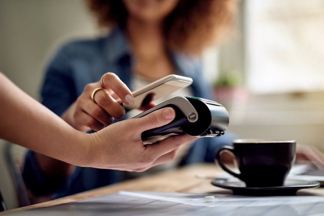 A person holds a smartphone up to a payment terminal to make a contactless payment at a table, with a cup of coffee in a quiet restaurant setting