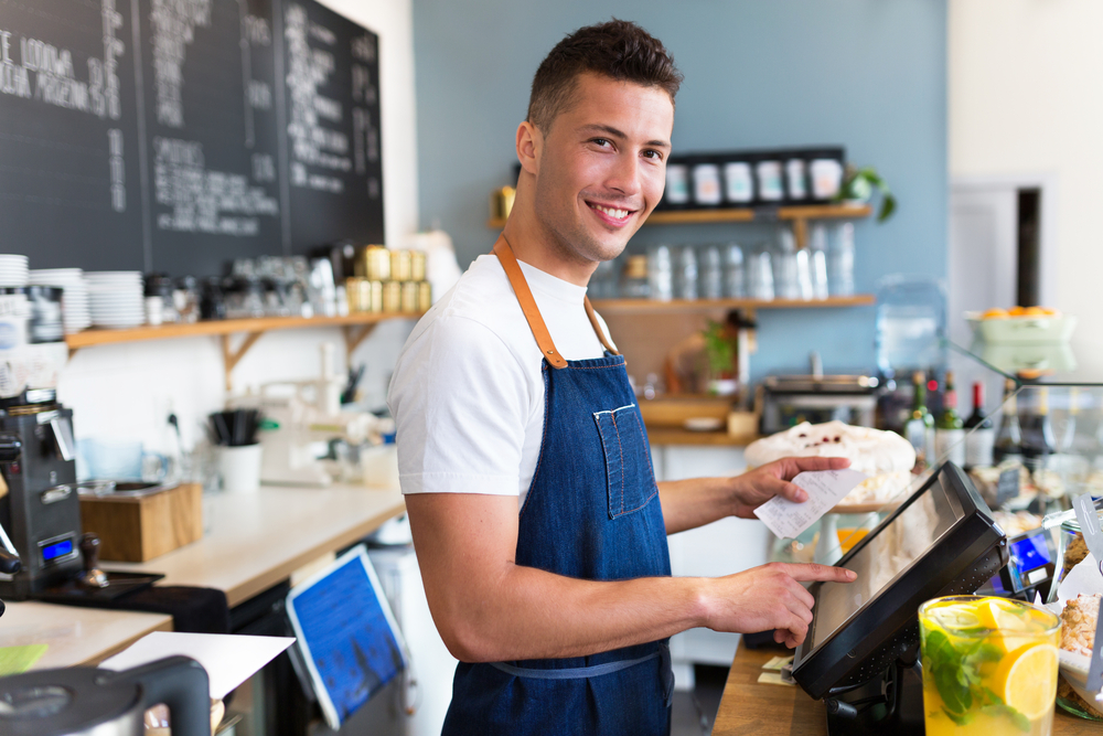 A café owner standing behind the register, smiling warmly at the camera while tapping on a screen and holding a receipt in his other hand