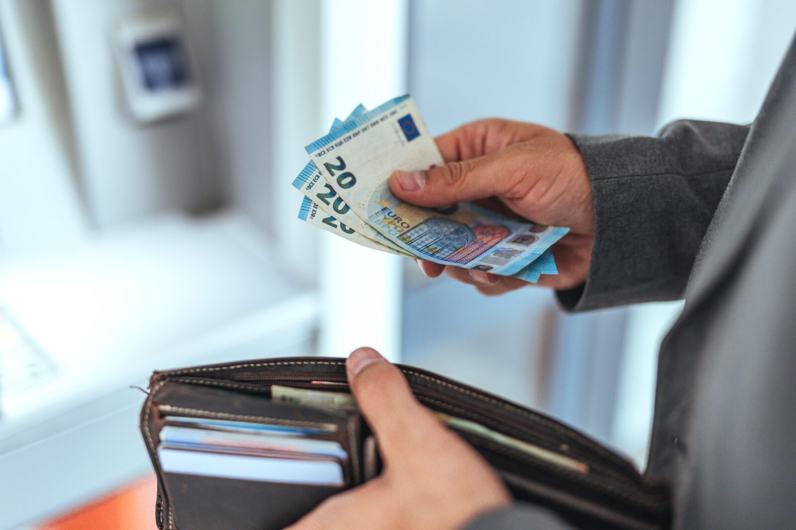 Image of a person standing in front of an ATM, holding three 20-euro bills in their right hand and a brown wallet containing cards in their left hand